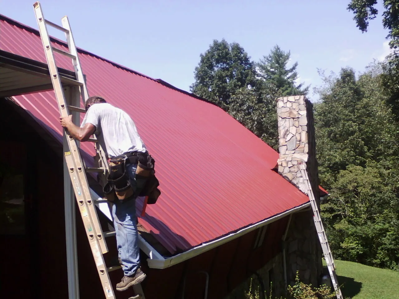 Roofer on a ladder beside a red metal roof and stone chimney — Soffit & Fascia Repair by Roofing Brothers in Cresskill